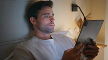 Yatakta tablet gibi duran bir adam rahat bir akşam tatili çekimleri yapıyor. Tıraşsız adam, sakin ev gecesinde dijital ekrana odaklanıyor. Yatak odasındaki bilgisayarda sosyal medyada gezinen sakin erkek 