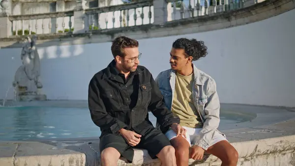 stock image Relaxed lgbtq partners sitting town fountain edge talking flirting together. Multiracial homosexuals relaxing in sunny weekend outdoors. Happy lovely gay couple chilling enjoying old city architecture