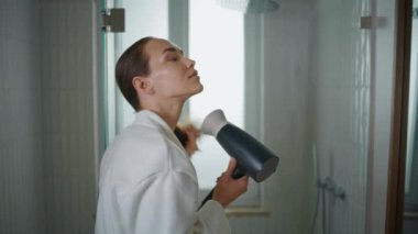 Girl blowing hair home after bath closeup. Happy lady getting ready in bathrobe. Attractive smiling woman looking mirror use hairdryer for styling in light bathroom. Beauty routine wellness in morning