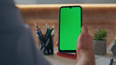 Kapalı alandaki telefon ekranını elle dinleyen bir adam. Bilinmeyen gevşek adam Chromakey akıllı telefonuna dokunuyor ofis yerinde iş e-postalarını kontrol ediyor. Elinde yeşil ekran cep telefonu zumlaması tutan isimsiz serbest çalışan 