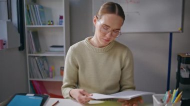 Ciddi bir kadın sınıf ödevlerini tek başına yakından kontrol ediyor. Gözlüklü öğretmen kadın elinde kalemle işyerinde notlar tutuyor. Yorgun eğitimci kabinede mola vererek defteri kapatıyor