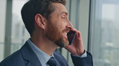 Büyük vitrinlerde cep telefonuyla konuşan çok mutlu bir patron. Mutlu finansçı telefonda pazarlık yapıyor şehir manzarasında neşeli sohbetler yapıyor. Gülümseyen müdür cam ofiste başarıya seviniyor 