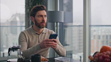 Düşünceli bir adam elinde cep telefonuyla büyük pencerelerin önünde kahvaltıdan zevk alıyor. Lüks bir dairede oturmuş akıllı telefon mesajı okuyan gülümseyen adam. Mutlu esmer sörfçüler evde internet gezintisi yapıyor.