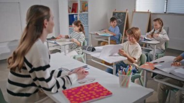 Grup öğrencileri okul sınıfını kapatıyorlar. Genç bayan öğretmen derste sorular soruyor. Aktif sınıf arkadaşları sıralarında gülümsüyor. Dost canlısı bayan öğretmen sınıfta çocuklarla konuşuyor. 