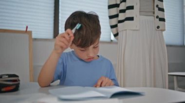 Sınıfta çalışan düşünceli şirin bir çocuk yakın çekim masasında oturuyor. Anaokulu öğrencisi, elinde kalem tutarak egzersiz kitabına yazmış. Zeki öğrenci cevapları içeride öğreniyor. 