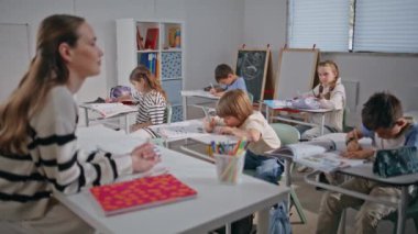 Bayan öğretmen ışık sınıfı yakın planında dersi açıklıyor. Öğrencilerle konuşan genç bir öğretmen. İlköğretim sınıfında not defteri arayan okul çocuklarına odaklanmış. Sınıf çalışması yapan küçük öğrenciler.
