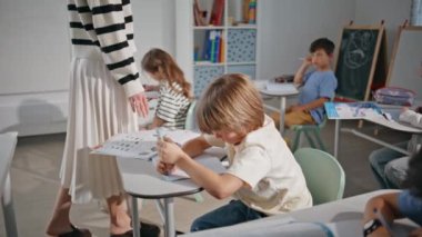 Okul sınıfında not defteri yazma sınavı yapan çocuklar grubu yakın çekim yapıyor. Öğrencilerle sıraların arasında yürüyen tanınmayan bir öğretmen. Sınıfta öğrencilere bakan tanınmamış bayan öğretmen.