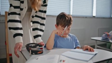 Sınıftaki öğrenciye yakın çekimde yardım eden genç bayan. Bayan öğretmen okul çocuğuyla konuşuyor. Sınıf çalışmalarını kontrol etmek için. Gülümseyen öğretmen kabinede öğrenciyle konuşuyor. Eğitim kavramı