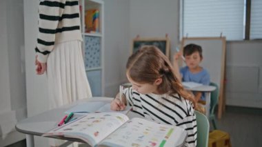 İlköğretim sınıfında not defterine yazma alıştırmaları yapan zeki kız. Küçükler sınıfında özenle çalışan tatlı bir çocuk dinleme dersi. Öğretmenler kabinede kız öğrencilere yardım ediyor.
