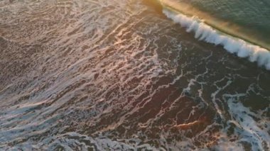 Drone manzaralı boş kumlu sahil. Gün batımında çarpıcı fırtınalı okyanus. Güçlü köpüklü dalgalar altın deniz kıyısını yavaş çekimde yıkıyor. Manzaralı vahşi deniz suyu güzel sahil yaz gününde akıyor. 