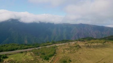 Virajlı yolu ve sakin atmosferi olan manzaralı dağlar. Hava manzaralı yeşil vadi ve beyaz kabarık bulutların altındaki yemyeşil tepeler. Asfalt yol, canlı yeşil yamaç boyunca uzanıyor. Huzurlu manzara