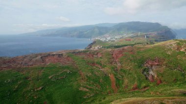 Panorama uzak tropikal ada hava manzaralı. Yaz sahilindeki sahil kasaba binaları. Bulutlu gökyüzünün altında uçan yeşil volkanik alanlar. Mavi okyanus huzurlu kıyı şeridini yıkıyor. Manzaralı tatil yeri