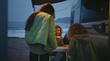 Mutlu arkadaşlar kasvetli okyanus kıyısındaki karavanda toplanıyorlar. Güler yüzlü bir grup kadın alacakaranlıkta karavanın terasında birlikte oturarak konuşuyor. Neşeli bayanlar deniz kıyısında dostça takılmaktan hoşlanırlar..