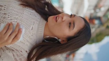 Turist kadın, şehir manzarasında kahve fincanıyla selfie çekiyor. Havalı Asyalı kız akıllı telefon kullanıyor. Hafta sonu dikey çekimde fotoğraf çekiyor. Gülümseyen esmer, güneşli caddede poz veriyor.