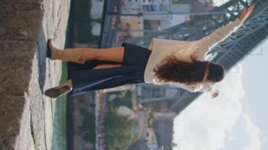 Şehir manzaralı nehir kıyısında yürüyen mutlu turistler. Neşeli bir kadın kollarını kaldırarak sette geziniyor. Tasasız, güzel bir kadın yaz gezintisinin keyfini çıkarıyor. Dikey dönüş videosu. Seyahat kavramı