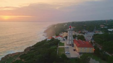 Yeşil sahildeki deniz feneri manzaralı küçük bir köyün üzerinde şaşırtıcı bir gün doğumu. Huzurlu okyanus yüzeyinde sabah gökyüzü köpüklü dalgalar dalgalarla dalgalı sahil şeridini süper yavaş çekimde yıkıyor..