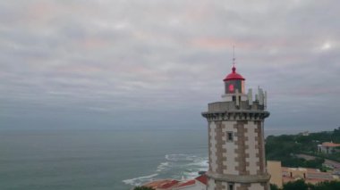 Deniz feneri insansız hava aracı çekiminde dramatik bir gökyüzü. Derin mavi okyanus bulutlu ufuk çizgisinin altında engebeli kıyı şeridini çok yavaş çekimde yıkıyor. Deniz feneri, bulutlu bir akşamda deniz taşımacılığı için yanıp sönüyor.