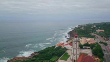 İnsansız hava aracı engebeli okyanus kıyısına akşam bulutlu gökyüzünün altına yerleştirilmiş kıyı köyünü vurdu. Deniz feneri yön bulma sinyali için kıyı şeridinde duruyor. Sert sahilin köpüklü dalgaları süper yavaş çekimde çarpıyor..