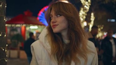 Gece karanlığında Noel fuarında eğlenen kızlar yakın çekim yapıyor. Aydınlanmış eğlence parkında tek başına kameraya bakan gülümseyen kadın. Tatil akşamı sokakta poz veren neşeli kadın model..