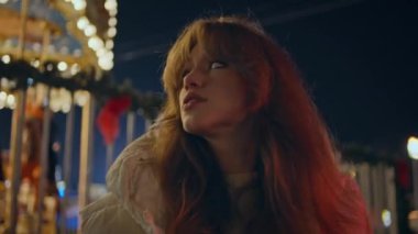 Tasasız kız Noel fuarı gecesinde atlıkarınca gibi görünüyor. Rahat kadın, parlak ışıklarla festival alanının tadını çıkarıyor. Havalı bayan hipster hafta sonu karanlık eğlence parkında tek başına dinleniyor..