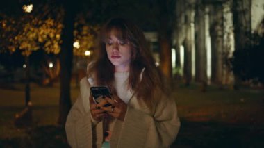 Millennial cep telefonunu şehir parkına yakın çekimde tutuyor. Sokak lambalarıyla aydınlatılmış ciddi esmer mesajlı akıllı telefon ayakta duran cadde. Karanlık şehirde telefon bekleyen çekici bir kadın..