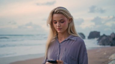 Gülümseyen kız Rocky Shore 'daki yakın çekimde SMS' leri kontrol ediyor. Günbatımı plajında rahat turist mesajları yazan akıllı telefonlar. Neşeli, çekici sarışın okyanus adasında cep telefonu iletişiminin keyfini çıkarıyor.