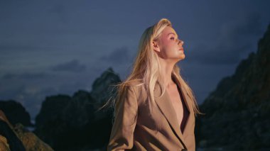 Bulutlu gökyüzünde kadın silueti akşam kumsalı. Kayalık Kıyı 'da seksi modelin üzerinde yanıp sönen ışık. Kendinden emin, baştan çıkarıcı sarışın, karanlıkta kaybolan bir kamera gibi. Bayan çıplak göğüslü şık bir takım elbise giyiyor.