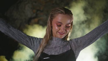 Duman bombası bulutu içinde dans eden mutlu kadın. Çekici kız projektör ışığında müzik dinlemekten zevk alıyor. Neşeli ve çekici bir bayan, karanlık arka planda festivale ayak uyduruyor.