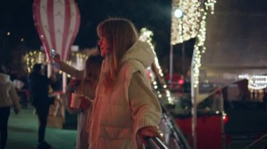 Gece festival fuarında elinde sıcak içecekle rahatlayan bir kız. Aydınlatılmış Noel ışıklarıyla çevrili karanlık sokakta duran rahat kadın. İyi tatiller. Aydınlatılmış parkta güzel bir hafta sonu..