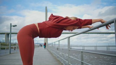 Kentsel koyda spor yapan atletik bir kadın. Sporcu kadının vücut bükme egzersizi yapmadan önce ısınması. Aktif sağlıklı kız koşucu deniz kıyısında spor kaslarını esnetiyor. Fitness rutini
