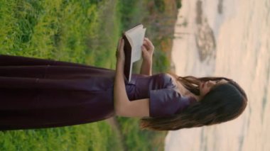 Romantik, uzun saçlı, kitap arayan, okyanus bulutlu bir günde sayfaları dikey çeviren bir kadın. Çekici bayan, yeşil çimen kıyısında tek başına kitap okuyor. Dalgın esmer, keyfine bak