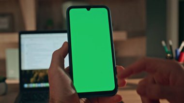 Ev işyerindeki krom anahtarlı cep telefonunu dizüstü bilgisayarla takip eden bir adam. Yeşil ekran akıllı telefona dokunan tanınmayan serbest çalışan. İş adamı elinde model cep telefonu mesajı tutuyor.