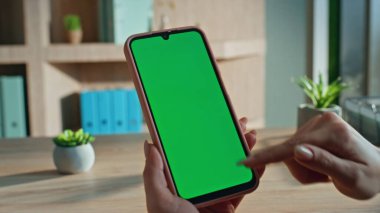 Krom tuşlu akıllı telefon araklayan serbest çalışan bir kız ev ofisinden online alışveriş yapıyor. Bilinmeyen iş kadını elleri dairede yeşil ekran cep telefonu tutuyor. Yönetici model telefona dokunuyor