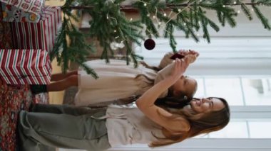 Anne kızı evde dikey videoda Noel ağacını süslüyor. Gülümseyen anne çocuk oturma odasındaki Xmas ormanına oyuncak koyuyor. Sevimli aile yeni yıl kutlaması için hazırlanıyor. Kış tatili 