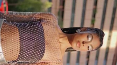 Kalabalık caddeye dikey odaklı yakın çekim yapan çekici bir moda kamerası. Şehirde poz veren şık kıyafetler giyen Afro-Amerikan kız portresi. Siyah örgülü esmer kadın tek başına ayakta duruyor.
