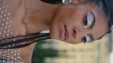 Gülümseyen kadın dikey videoda makyaj aynasını kontrol ediyor. Afrika kökenli Amerikalı moda tutkunu küpelere dokunup yaz akşamı sokağına bakıyor. Moda insan yaşam tarzı konsepti