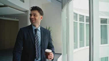 Rahatlamış girişimci elinde kahve paketiyle ofis koridorunun yanında yürürken panoramik pencereleri kapatıyor. Kendine güvenen işadamı cep telefonunu kontrol ederken gülümsüyor. Mutlu Ceo görünümlü akıllı telefon bardağı içeceği.