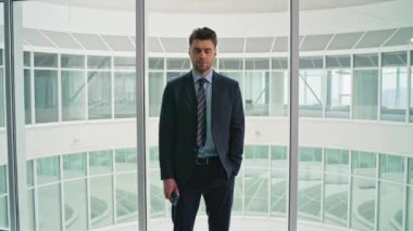 Ofis panoramik pencerelerinde duran zarif iş adamının portresi yakınlaştır. Ciddi bir işletme yöneticisi kameraya akıllı telefon tutuyor. Modern şirket koridorunda takım elbiseli bir erkek..