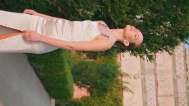 Doğayı rahatlatan zarif bir model. Yumuşak güneş ışığı altında yüzünü yakınlaştırıyor. Yeşil park yolunda dikey vurulan beyaz elbiseli rahat bir kadın. Tasasız kız güzel bahçe manzarasının tadını çıkarıyor.