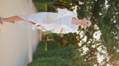 Rüzgarda sallanan beyaz eşarbıyla yeşil yolda yürüyen sakin bir kadın. Gülümseyen zarif bir bayan dikey dikey antik heykellerle patika parkına gidiyor. Rahatlamış kaygısız kız doğada geziniyor..