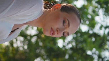 Yeşil Park 'ın doğasına poz veren büyüleyici kadın kameraya yakından bakıyor. Ağaçların altında dikey güneş ışığı videosunun keyfini çıkaran muhteşem bir kadın. Güzel manken gözlerini kapatıyor parkta dinleniyor.