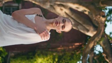 Parkta beyaz kıyafetlerle poz veren şehvetli bir model. Yüksek yeşil ağaçların yanında dikey çekim yapan genç ve ciddi bir bayan. Gevşemiş hayalperest kadın güneşli yaz doğasının tadını çıkarıyor..