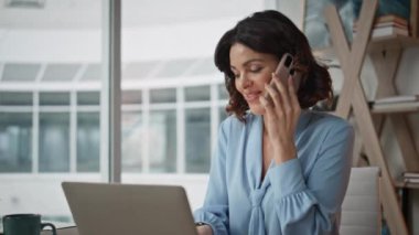 Ofis masasında dizüstü bilgisayarla cep telefonundan konuşan mutlu bir çalışan. Gülümseyen kadın yönetici modern işyerinde cep telefonu sohbetinde bulundu. İş kadını işi tartışıyor ve tatmin oluyor.