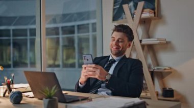Modern ofis koltuğunda oturan neşeli profesyonel cep telefonu akşam kapanışı. Mutlu yönetici akıllı telefondan mesaj atıyor. Gülümseyen iş adamı cep telefonunda sosyal medyaya göz atıyor..