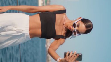 Bayan turist, Sunny rıhtımındaki dikey merkezde akıllı telefondan selfie çekiyor. Güneş gözlüklü kadın rıhtımda duran cep telefonu kamerasına bakıyor. Sosyetik kız deniz manzarasının önünde fotoğraf çekiyor..