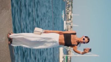 Deniz rıhtımında cep telefonunu dikey tutan gülümseyen kadın blogu. Lüks yatlarla deniz manzarası kaydeden güzel bir kız. Güneş gözlüklü güzel bayan deniz manzaralı içeriği çekiyor.