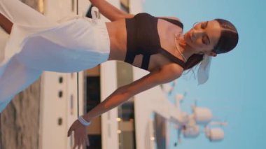 Rıhtımda poz veren modacı kız kameraya güvenle dikey odaklı bakıyor. Beyaz yatın önünde iskelede duran genç bir kadın. Gülümseyen bayan turist yaz tatilinde deniz kenarında dinleniyor.