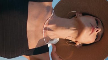 Mavi gökyüzüne dikey yakın plan pozu veren zarif kadın hasır şapka. Moda bir başlıkla yaz seti kenarında duran şık modelin portresi. Şatafatlı zengin kız güneşli doğada kameraya bakıyor..