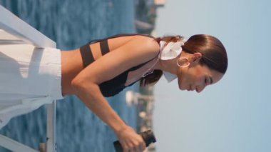 Dürbünlü kadın yaz gezisinde deniz güzelini dikey izliyor. Güneşli sahil kıyısındaki mutlu çekici kız turist güzel deniz manzarasının tadını çıkarıyor. Moda tebessümlü esmer bayan kamera kıyıda poz veriyor.