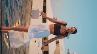 Şık, modaya uygun, dikey kostüm videosuyla deniz rıhtımında duran bir model. Lüks yatın önünde poz veren güzel, kendine güvenen bir kadın limana demirlemiş. Şık kız yazın dinleniyor. Tasasız tatil